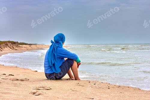 Preview: A Woman In A Blue Scarf And A Sweater On Her Head Is Sitting On The Beach By The Sea
