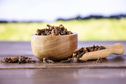 Preview: Lot Of Whole Small Dried Cloves Spice Scoop With Wooden Bowl With Green Wheat Field In Background