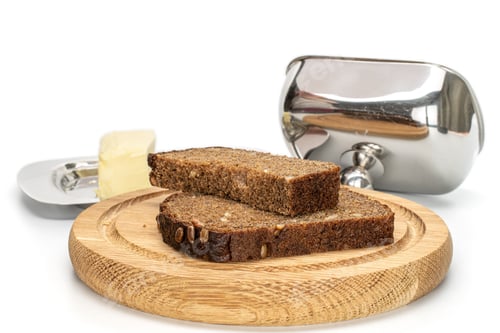 Preview: Group Of Two Slices Of Fresh Baked Dark Bread On Bamboo Plate With Butter Dish Isolated On White