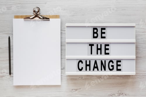 Preview: 'Be The Change' Words On A Lightbox, Clipboard With Blank Sheet Of Paper On A White Wooden Surface