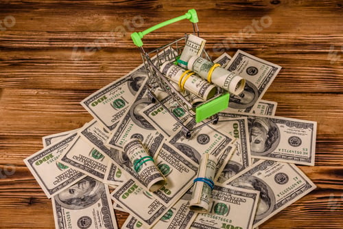 Preview: Small Shopping Cart With Rolled Up One Hundred Dollar Banknotes On Wooden Background