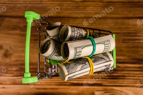 Preview: Small Shopping Cart With Rolled Up One Hundred Dollar Banknotes On Wooden Background. Top View
