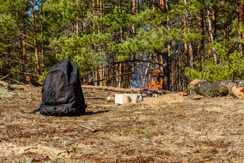 Preview: Backpack On A Ground In Coniferous Forest. Bonfire On A Background