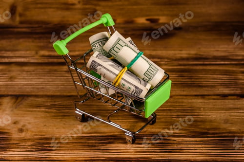 Preview: Small Shopping Cart With Rolled Up One Hundred Dollar Banknotes On Wooden Background