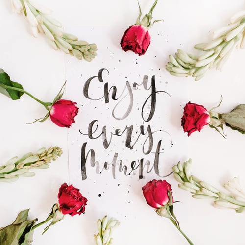 Preview: Inspirational Quote "Enjoy Every Moment" Written In Calligraphic Style On Paper With Red Roses