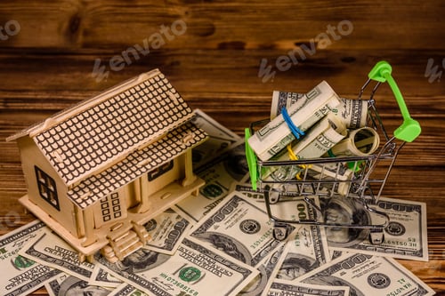 Preview: Small Shopping Cart With Rolled Up One Hundred Dollar Banknotes And Plywood Model Of The House On