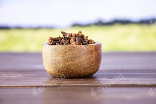 Preview: Lot Of Whole Small Dried Cloves Spice With Wooden Bowl With Green Wheat Field In Background