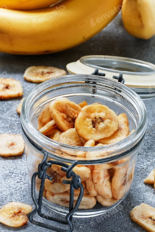 Preview: Banana Chips, Dehydrated Slices Of Fresh Ripe Bananas In A Glass Jar On A Grey Concrete Background