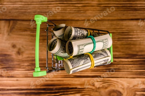 Preview: Small Shopping Cart With Rolled Up One Hundred Dollar Banknotes On Wooden Background. Top View