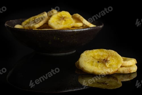 Preview: Lot Of Slices Of Sweet Yellow Dry Banana In Dark Ceramic Bowl Isolated On Black Glass