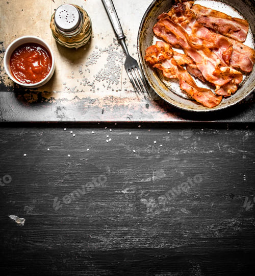 Preview: Fried Bacon In A Frying Pan With The Sauce. On A Black Wooden Background.