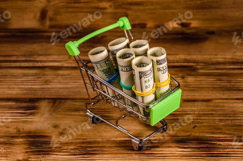 Preview: Small Shopping Cart With Rolled Up One Hundred Dollar Banknotes On Wooden Background