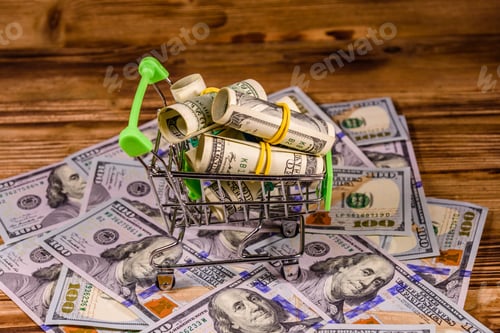 Preview: Small Shopping Cart With Rolled Up One Hundred Dollar Banknotes On Wooden Background