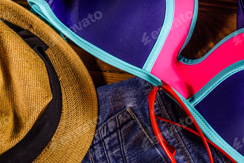 Preview: Female Swimming Suit, Denim Shorts, Straw Hat And Eyeglasses On Rustic Wooden Table. Top View