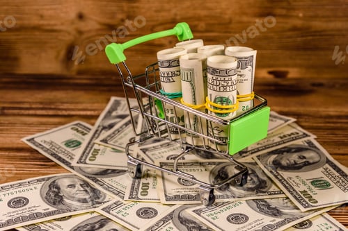 Preview: Small Shopping Cart With Rolled Up One Hundred Dollar Banknotes On Wooden Background