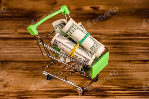 Preview: Small Shopping Cart With Rolled Up One Hundred Dollar Banknotes On Wooden Background