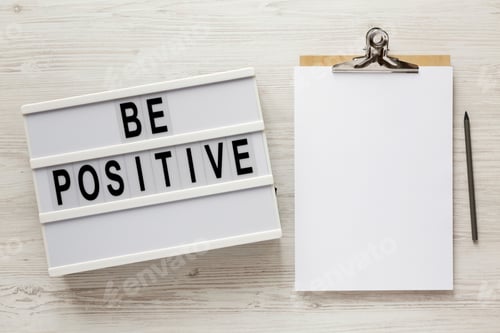 Preview: 'Be Positive' Words On A Lightbox, Clipboard With Blank Sheet Of Paper On A White Wooden Surface