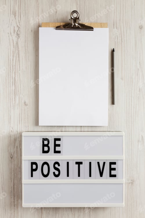 Preview: 'Be Positive' Words On A Lightbox, Clipboard With Blank Sheet Of Paper On A White Wooden Surface