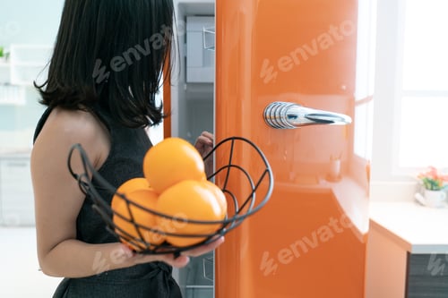 Preview: Woman Open Refrigerator Door In The Kitchen. Orange In Hand.