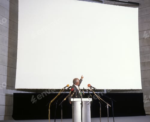 Preview: Businessman At Podium Pointing At Large Projection Screen
