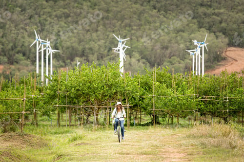 Preview: Woman Riding Bicycle In Field With Wind Turbines In Background