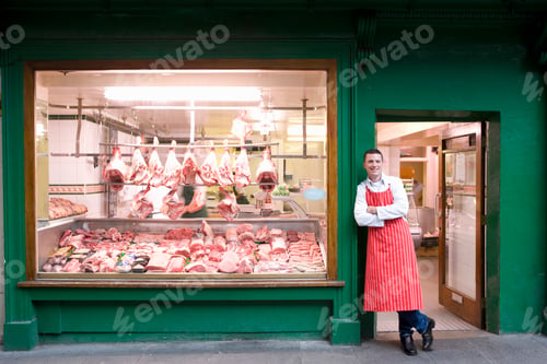 Preview: A Wide Shot Of A Young Smiling Butcher In Red Apron Leaning Against Butcher Shop Doorway Next To