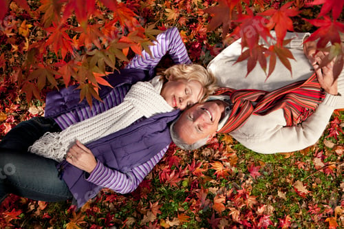 Preview: A Top Angle Shot Of A Happy Senior Couple Laying On The Grass With Red Autumn Leaves In Foreground.
