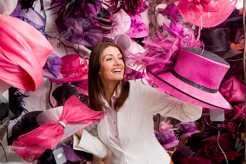 Vista previa: A Smiling Woman Looking At Different Ornate Feathered Hats In A Shop.