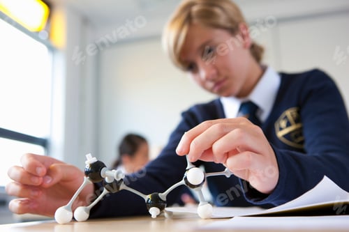 Preview: A Close Up Shot Of A Young Blonde Girl In School Uniform Looking Down At An Atom Model On Her Desk