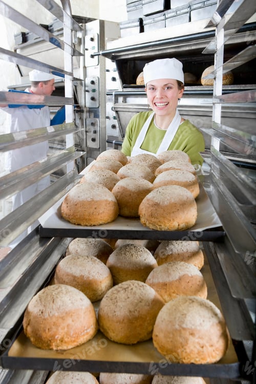 Preview: A Portrait Shot Of A Happy Female Baker Putting A Pan Of Fresh Loaves Of Bread In A Rack.