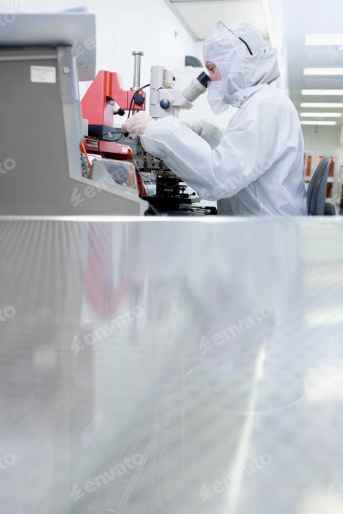Preview: A Distant Vertical View Of A Scientist In Clean Suit Examining The Silicon Wafer Under A Microscope