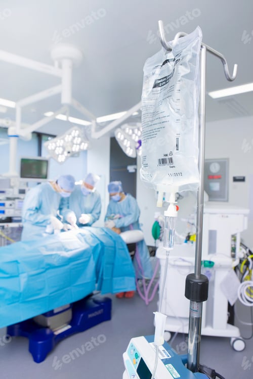 Preview: A Vertical View Of An Operating Room With Iv Bag Hanging In The Foreground While A Group Of