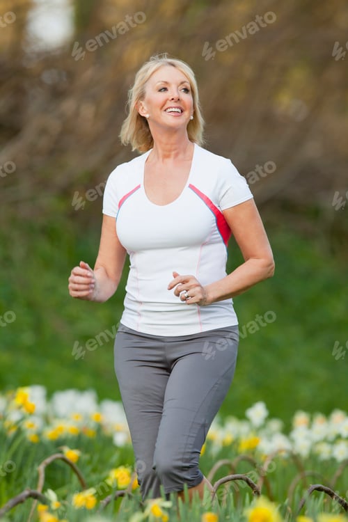 Preview: Low Angle Shot Of A Smiling Senior Woman In Activewear Jogging By A Daffodil Field On An Overcast