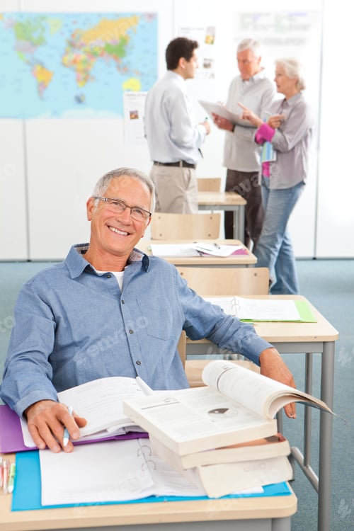 Preview: A Portrait Shot Of A Senior Man With Gray Hair Smiling At Camera And Sitting On A Desk With A Pile