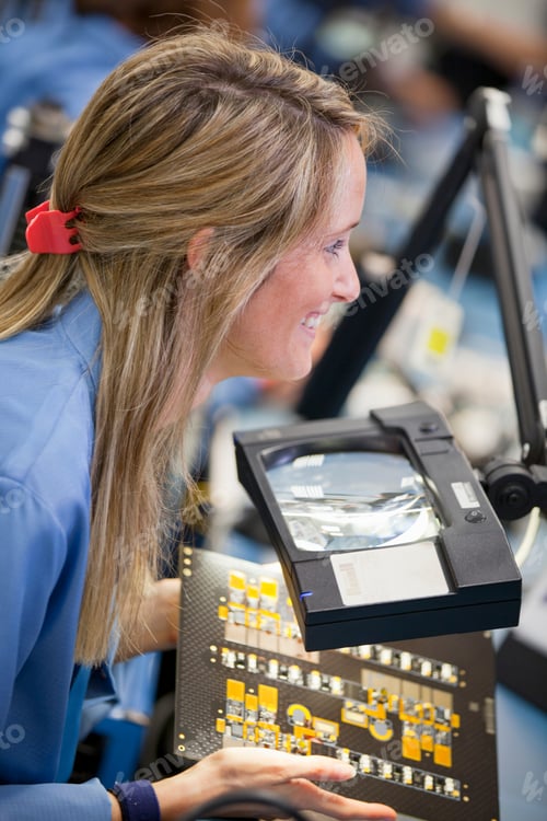 Preview: Vertical Profile Shot Of A Smiling Female Technician At Her Desk Inspecting A Printed Circuit Board