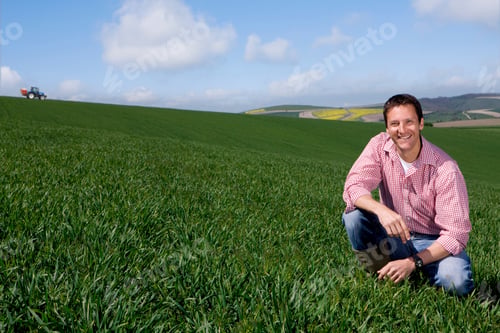 Preview: A Farmer Kneeled Down In A Wheat Field And Smiling At The Camera With A Tractor In Background.