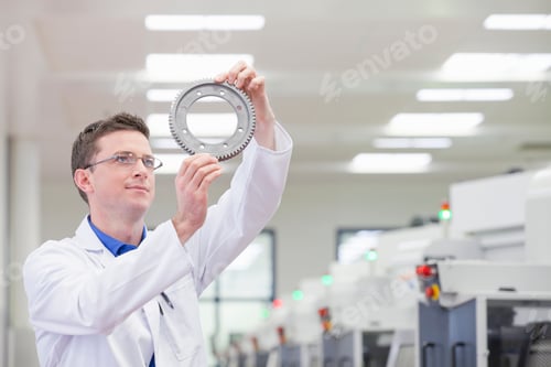 Preview: Horizontal Shot Of A Scientist Holding Up And Examining A Gear Wheel In A Manufacturing Plant With