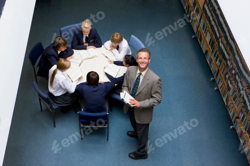 Preview: A High Angle Medium Shot Of A Teacher Smiling At Camera And His Students Reading Books At A Round