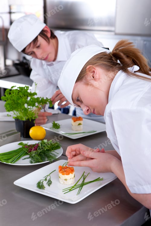 Preview: A Close Up Vertical Shot Of Two Young Trainee Chefs Garnishing Desert In A Kitchen.