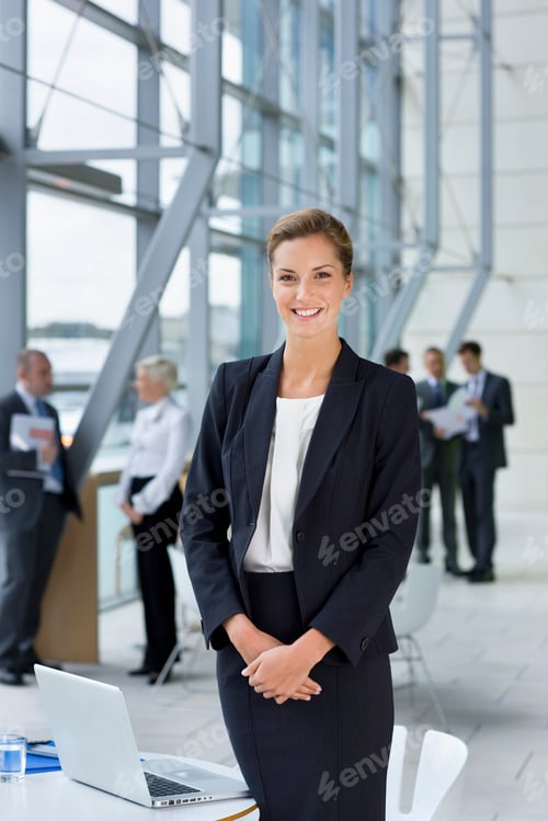 Preview: A Portrait Of A Smiling Businesswoman In A Formal Suit While Standing Next To A Laptop On A Table