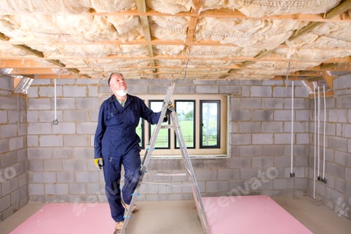 Preview: A Man In Coveralls Standing On A Ladder And Looking Up At Ceiling Insulation In An Attic.