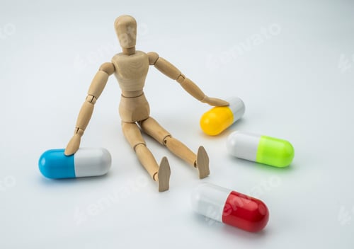Preview: Articulated Wooden Doll Seated Along With Capsules Of Different Colors, Given Shelter On White Fund