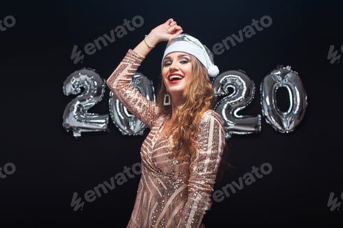 Preview: Happy Young Woman In Santa Hat Having Fun Against Metallic 2020 Balloons On Black Background. New