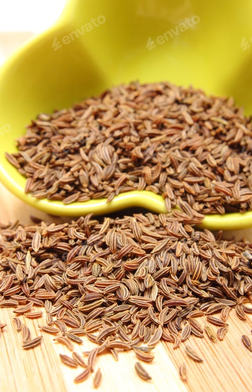 Preview: Closeup Of Fresh Brown Cumin Pouring Out Of Yellow Bowl On Wooden Cutting Board