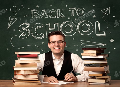 Visualização: Um jovem professor de óculos sentado na mesa da sala de aula com uma pilha de livros na frente do quadro-negro