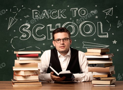 Visualização: Um jovem professor de óculos sentado na mesa da sala de aula com uma pilha de livros na frente do quadro-negro
