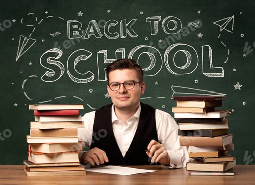 Visualização: Um jovem professor de óculos sentado na mesa da sala de aula com uma pilha de livros na frente do quadro-negro
