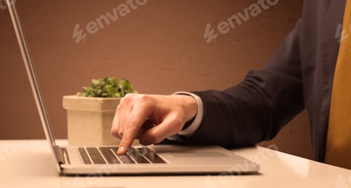 Preview: An Office Worker In Elegant Suit Sitting At Desk, Typing On Portable Laptop With Empty Brown Wall