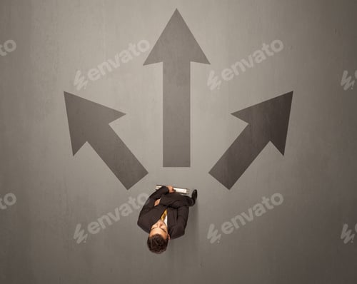 Preview: Young Contemplating Businessman Stands In Front Of Brown Arrows