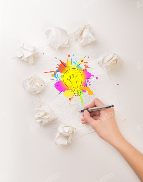 Preview: Female Hand Next To A Few Crumpled Paper Balls Drawing A Colorful Lightbulb
